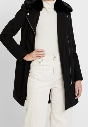 Zwarte wollen jas met een nepbontkraag, ritsluiting en gestructureerde vorm. Gepaard met crèmekleurige corduroy broek. Geweven stof zichtbaar.