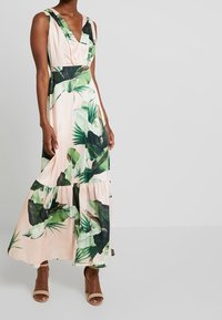Robe longue rose pâle à imprimé feuilles tropicales vertes ; encolure en V profonde, sans manches, ourlet à volants étagé. Texture de tissu lisse.