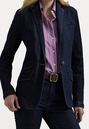 Kobieta nosząca ciemny jeansowy żakiet i spodnie dżinsowe, połączone z fioletową koszulą w paski zapinaną na guziki oraz brązowym paskiem z srebrną klamrą.