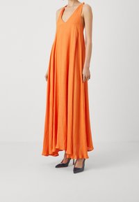 Robe maxi sans manches orange avec un décolleté en V, une silhouette fluide et un tissu texturé ; assortie à des escarpins bleus marine à bout pointu.