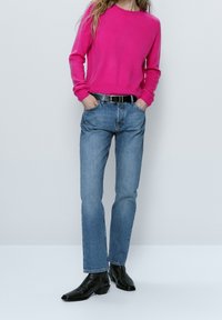 Pull fuchsia, jeans slim fit bleu, ceinture noire, bottes en cuir à la cheville. La tenue met en avant un contraste de couleurs vibrant et un design ajusté.