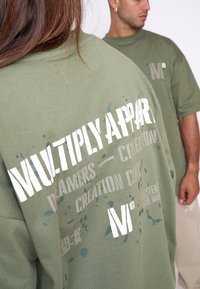 Camiseta de gran tamaño en color caqui con texto en blanco y azul y patrones de salpicaduras. Tejido de algodón suave, corte relajado, cuello redondo y mangas cortas.