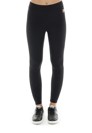 Une personne porte un legging noir taille haute et des baskets noires et blanches sur fond blanc.