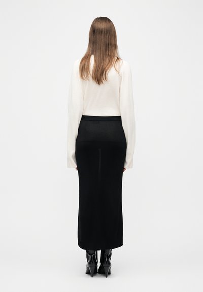 Γυναίκα με μακριά καστανά μαλλιά, φοράει λευκό μακρυμάνικο τοπ, μαύρη φούστα μέχρι τον αστράγαλο και μαύρα μπότες με τακούνι, στέκεται με την πλάτη προς το μέρος μας.