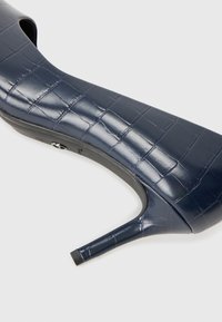 Scarpa nera con tacco alto e lucido, con un motivo a croco testurizzato. Presenta una punta appuntita e un sottile tacco a stiletto, progettata per una silhouette elegante.
