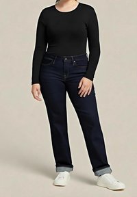 Personne portant un haut noir à manches longues, un jean bleu foncé retroussé et des baskets blanches, debout devant un fond uni.