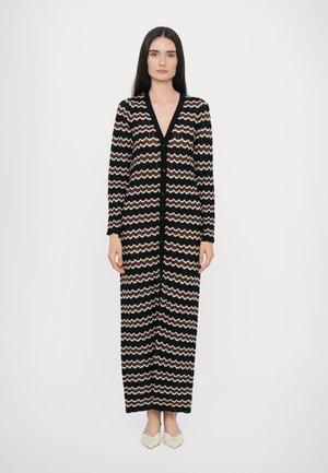 Lang zwarte cardigan met zigzagpatroon in beige en wit. Heeft een V-hals, lange mouwen en knopen aan de voorkant. Gladde textuur.
