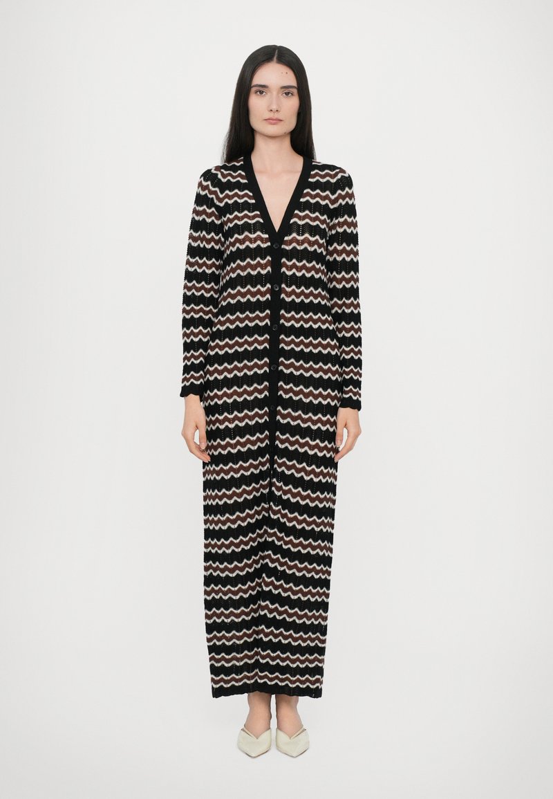 Lang zwarte cardigan met zigzagpatroon in beige en wit. Heeft een V-hals, lange mouwen en knopen aan de voorkant. Gladde textuur.
