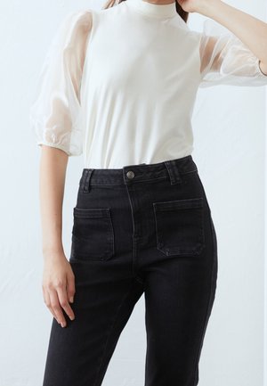 Weißes Oberteil mit durchsichtigen Puffärmeln, kombiniert mit hochtaillierten schwarzen Jeans mit zwei Gesäßtaschen. Einfaches, klares Design.