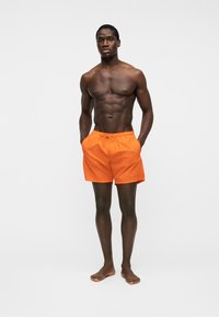 Jeune homme musclé debout pieds nus portant un short de bain orange vif, les mains dans les poches, sur un fond clair uni.