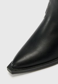 Bota de tobillo de cuero negra con punta afilada, diseño elegante y detalles mínimos. Cuenta con una suela plana y una construcción robusta.