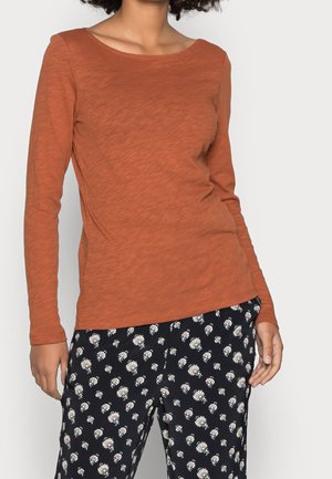 Person iført en rustfarvet langærmet top og sorte bukser med hvide blomsterprint, stående mod en lys baggrund.