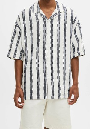 Mężczyzna w krótkim, białym koszuli na guziki z czarnymi pionowymi paskami i białych szortach, stojący na jednolitym tle.