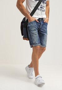 Shorts di denim blu chiaro, strappati con bordi sfilacciati, indossati con una maglietta bianca con grafica e sneaker bianche; portando una borsa a tracolla nera.
