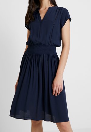 Woman wearing a navy blue short-sleeve dress with a V-neck, gathered waist, and knee-length flowy skirt on a white background.
