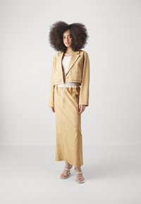 Veste beige cintrée avec deux poches avant portée sur un haut blanc et une jupe longue beige assortie. L'ensemble présente une taille à cordon de serrage et une texture lisse.