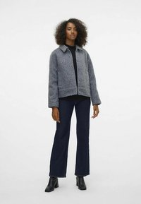 Pilka, tekstūruota trumpa striukė su apykakle, atvira priekyje ir ilgomis rankovėmis; derinama su tamsiais kelnėmis su platėjančiomis apačiomis ir auliniais batais.