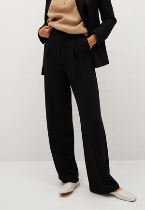 Kobieta ubrana w czarne spodnie o szerokich nogawkach, beżowy sweter z dzianiny, czarną marynarkę i białe czółenka na szpic, z rękami w kieszeniach, stojąca na jasnym tle.