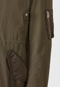 Olivgrön tyg med lock fäst med tryckknapp och en dragkedjeficka med silverdragkedja på ett plagg.