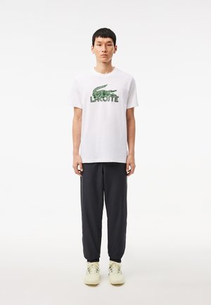 T-shirt bianca in cotone con logo del coccodrillo verde Lacoste, abbinata a pantaloni jogger grigio scuro e sneaker di colore chiaro. Design semplice e casual.
