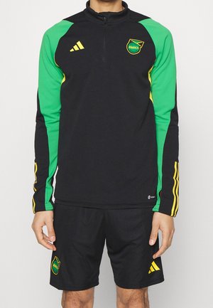 Man draagt een zwart-groen Jamaica Football Federation trainingsshirt en zwarte korte broek met gele Adidas-logo's en strepen.