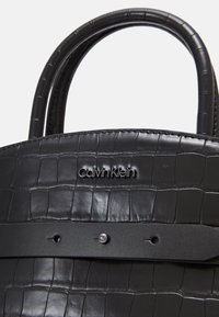 Černá taška z faux krokodýlí kůže s dvojitými uchy, vodorovným popruhem s kovovým knoflíkem "ck" a logem Calvin Klein na přední straně.