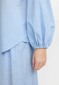 Top azzurro chiaro a righe con maniche voluminose e polsi arricciati; il tessuto ha una texture liscia con un dettaglio a orlo sovrapposto.