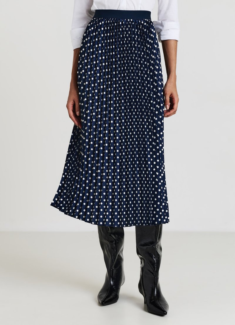 Jupe plissée bleu marine à pois blancs ; fabriquée en tissu léger. Elle possède une taille élastique et tombe jusqu'au milieu du mollet.