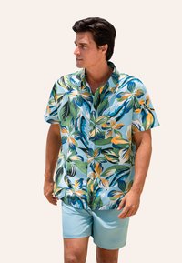 Koszula krótki rękaw, zapinana na guziki, o niebieskim tle i żywym tropikalnym wzorze kwiatowym w kolorach zielonym, żółtym i pomarańczowym; zestawiona z jasnoniebieskimi szortami.
