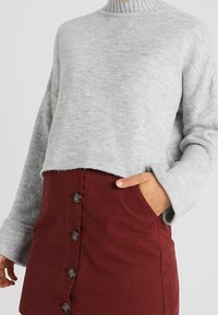 Grå, kort tröja med hög ribbad krage och vida ärmar, tillsammans med en vinröd kjol med knappar fram och sidofickor.