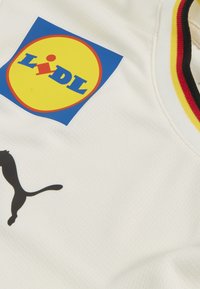 Jersey blanca con un logo de Lidl en azul y amarillo y un logo de Puma en negro. La tela tiene un patrón texturizado y acentos en negro, rojo y amarillo.