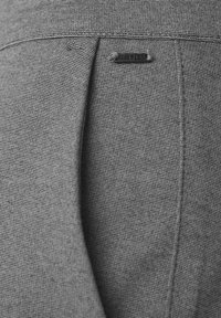Gros plan sur un pantalon en tissu gris texturé avec une petite étiquette noire rectangulaire cousue au-dessus d'une couture verticale.