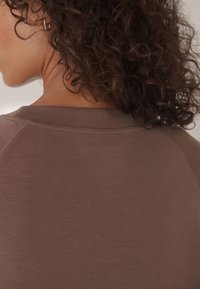 Maglietta marrone a maniche corte realizzata in tessuto liscio. Presenta una scollatura rotonda e un dettaglio testurizzato sul retro, con un logo discreto vicino al collo.