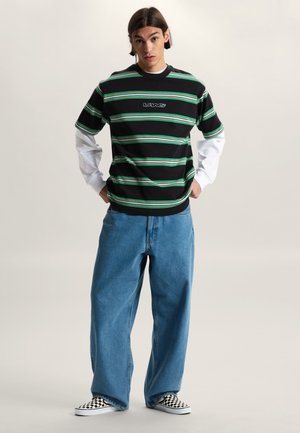 Jonge man staat met handen in zijn zakken, draagt een zwart-groen gestreept Vans-shirt over witte lange mouwen, wijde blauwe jeans en geruite slip-on schoenen.