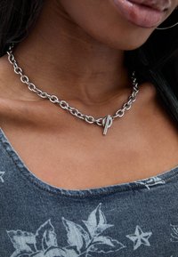 Ezüst lánc nyaklánc nagy, összekapcsolt karikákkal és egy két részből álló kapoccsal, amelyet egy kék virágmintás felsővel viselnek.