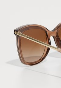 Ochelari de soare oversized, maro, cu lentile gradient. Prezintă un braț din metal de ton auriu și o montură din plastic transparent. Branding "Michael Kors" vizibil.