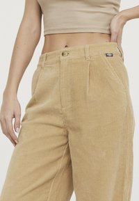 Pantalon en velours côtelé beige, présentant une texture à côtes verticales, une fermeture à un bouton et des poches latérales. Patch de marque notable présent.