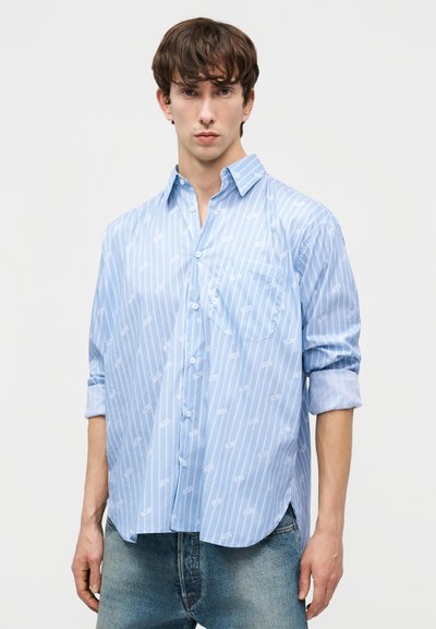 Jauns vīrietis, tērpies gaiši zilā svītrainā krekliņā ar uzvītu piedurkņu un gaiši zilās džinsu biksēs, stāv pret vienkāršu fonu.