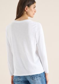 Camisa blanca de manga larga hecha de una tela ligera, con cuello redondo y un corte relajado, combinada con jeans azules.