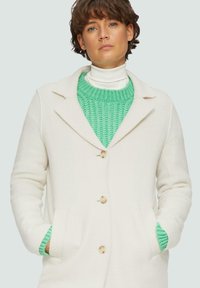Manteau blanc en mélange de laine avec un col à revers, des boutons à l'avant et des poches latérales. Superposé avec un pull en tricot vert menthe en dessous.