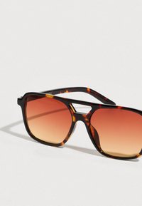 Γυαλιά ηλίου με τετράγωνα φακούς, βαθμιαία πορτοκαλί φακούς και χοντρό σκελετό με κομμένα στοιχεία στη γέφυρα.