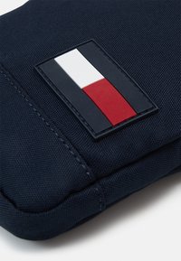 Roh černé látkové tašky s přišitým obdélníkovým výpletem zobrazujícím černé, bílé a červené svislé barevné bloky.