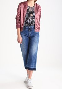 Rosa Satin-Bomberjacke über einem schwarzen Blumenshirt, kombiniert mit weit geschnittenen, hellblauen Jeans und weißen Sneakers. Lässiger, lockerer Schnitt.
