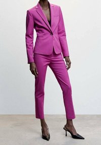 Blazer fuchsia sur mesure avec revers crantés et poche avant, associé à un pantalon slim assorti. Des escarpins noirs à bouts pointus complètent la tenue.