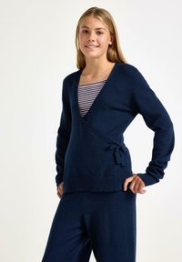 Ung kvinde iført en marineblå indviklet sweater med bånd og matchende bukser, smilende, mod en ensfarvet hvid baggrund.