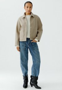 Beige Wollmischjacke mit Kragen, Druckknopfverschlüssen, zwei Fronttaschen, getragen über einem hellen Pullover und blauen oversized Jeans, kombiniert mit schwarzen Stiefeln.