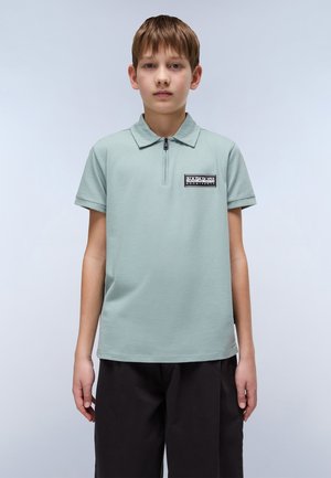 Polo shirt vert clair avec un col zippé à mi-hauteur, manches courtes et des détails de label noir. Porté avec un pantalon sombre. Texture lisse.