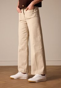 Crème-kleurige wijde jeans met een gladde textuur, voorzien van dubbele voorkeurszakken en een rits. Gecombineerd met witte sport schoenen.