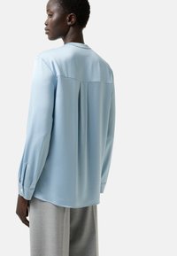 Blouse bleu clair en tissu lisse, dotée d'un pli au dos, de manches longues avec des poignets boutonnés et d'un ourlet arrondi.