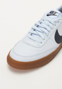 Gaiši zili ādas sporta apavi ar melnu Nike logotipu, plakaniem baltajiem šņorēm un teksturētu gumijas zoli. Klasiska apaļa purngalu forma.